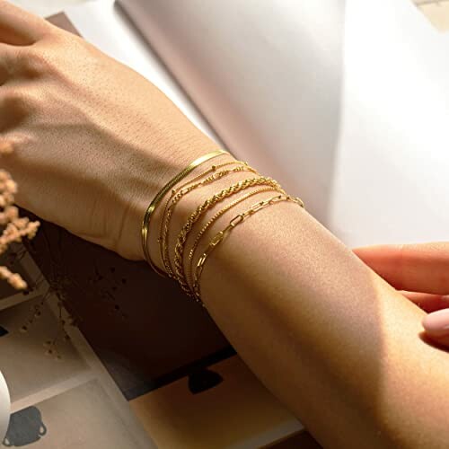 Person wearing multiple gold bracelets on wrist with open book in background.