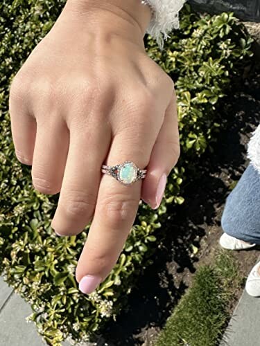Hand wearing an opal ring with greenery in the background.