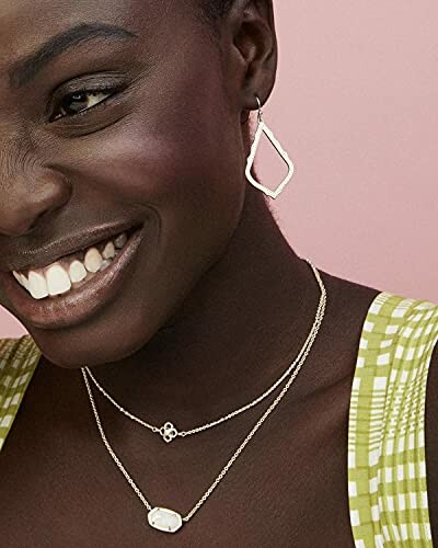 Woman smiling, wearing geometric earrings and layered necklaces.