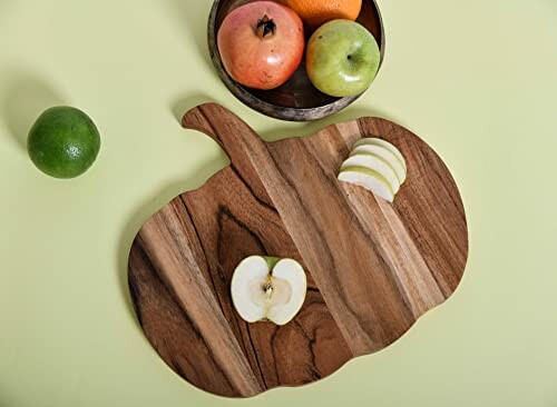 Pumpkin-shaped wooden cutting board with sliced apples and fruit bowl.
